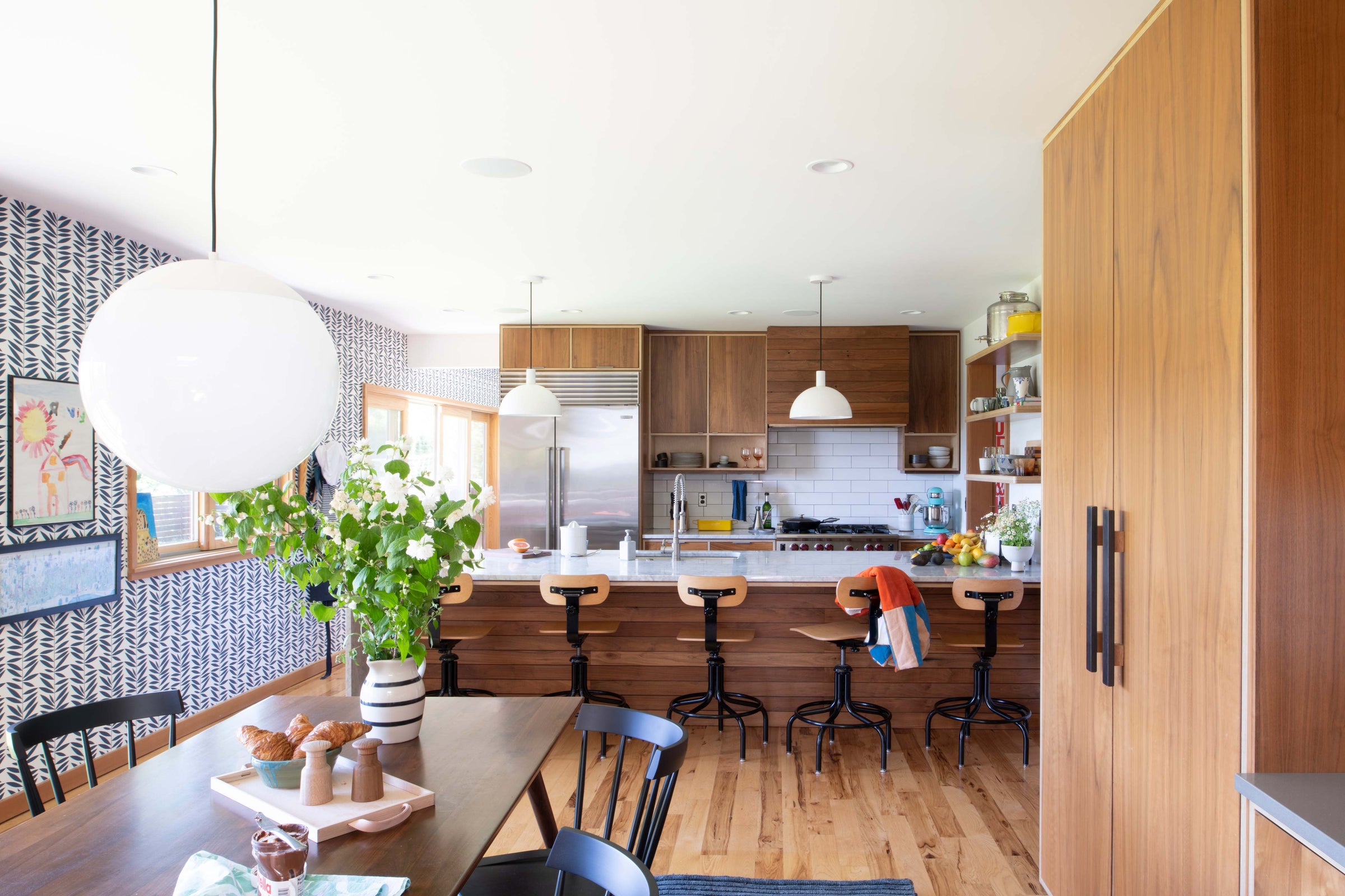 Sunlit kitchen and dining area with walnut cabinets, marble island, bar stools, patterned wallpaper, and vase of flowers