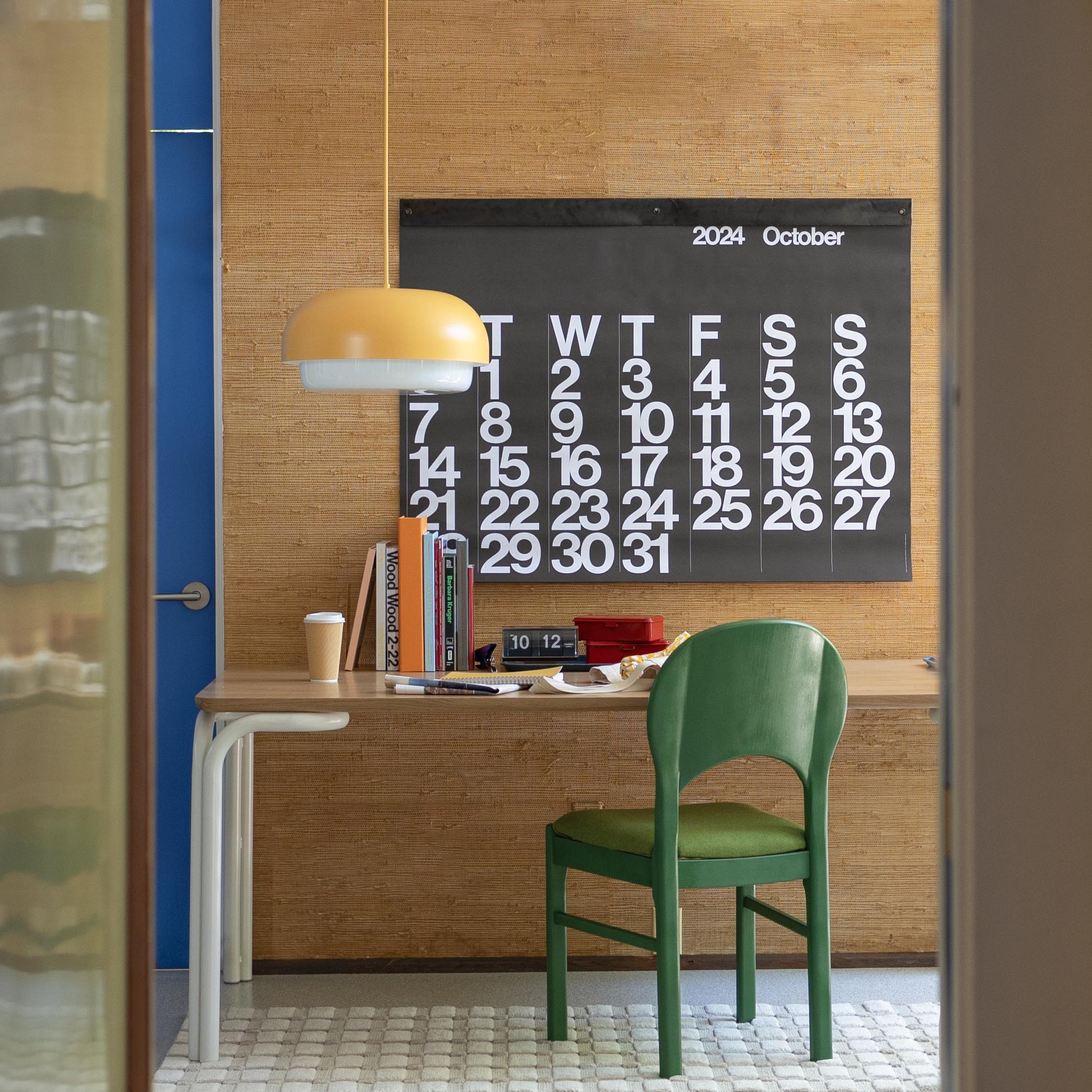 Workspace with green chair and wooden desk featuring a hanging yellow pendant and a large black wall calendar reading ‘2024 October’