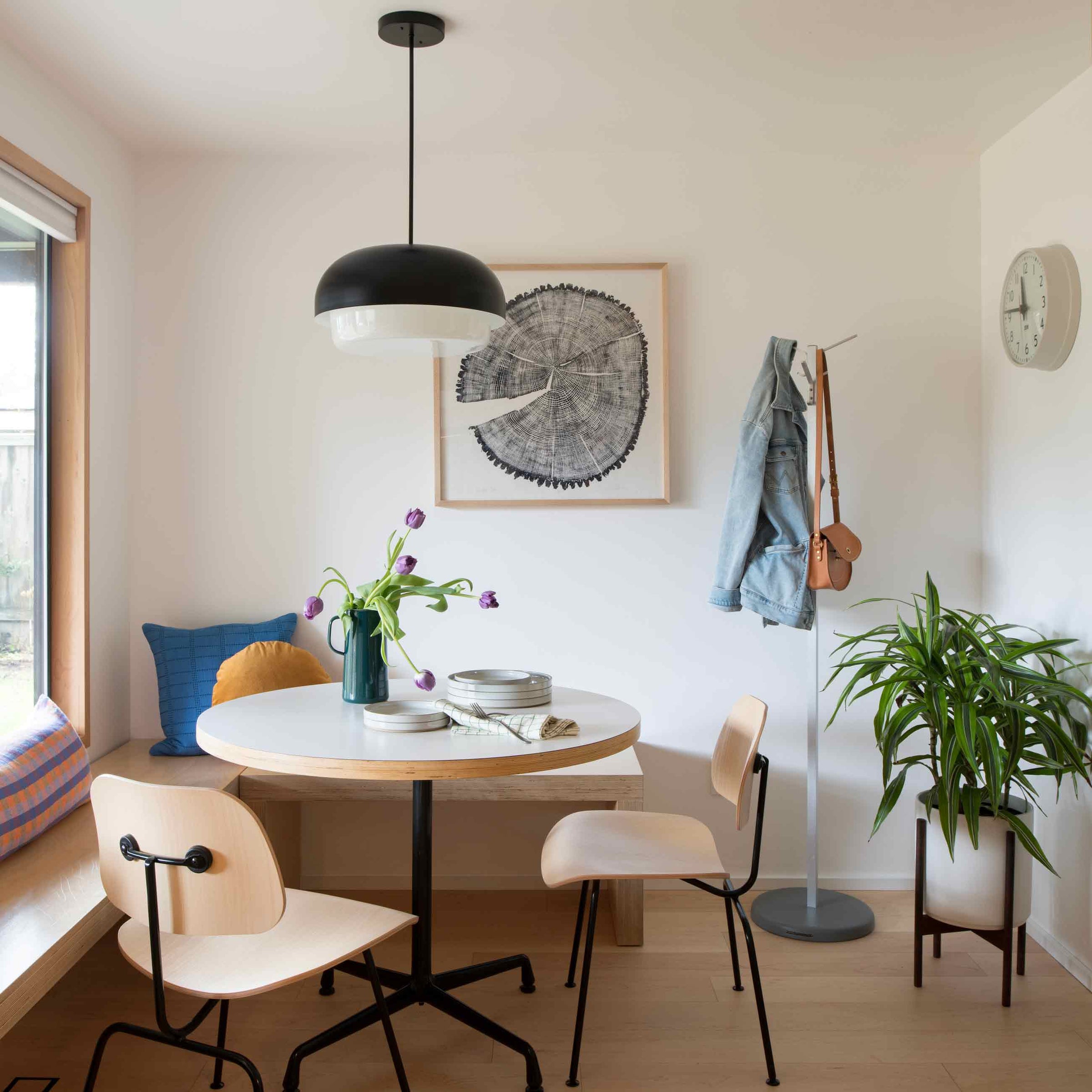 Bright minimalist dining nook with round white table, black dome pendant light, purple tulips in a green pitcher, light wood chairs, coat stand with denim jacket, and potted plant