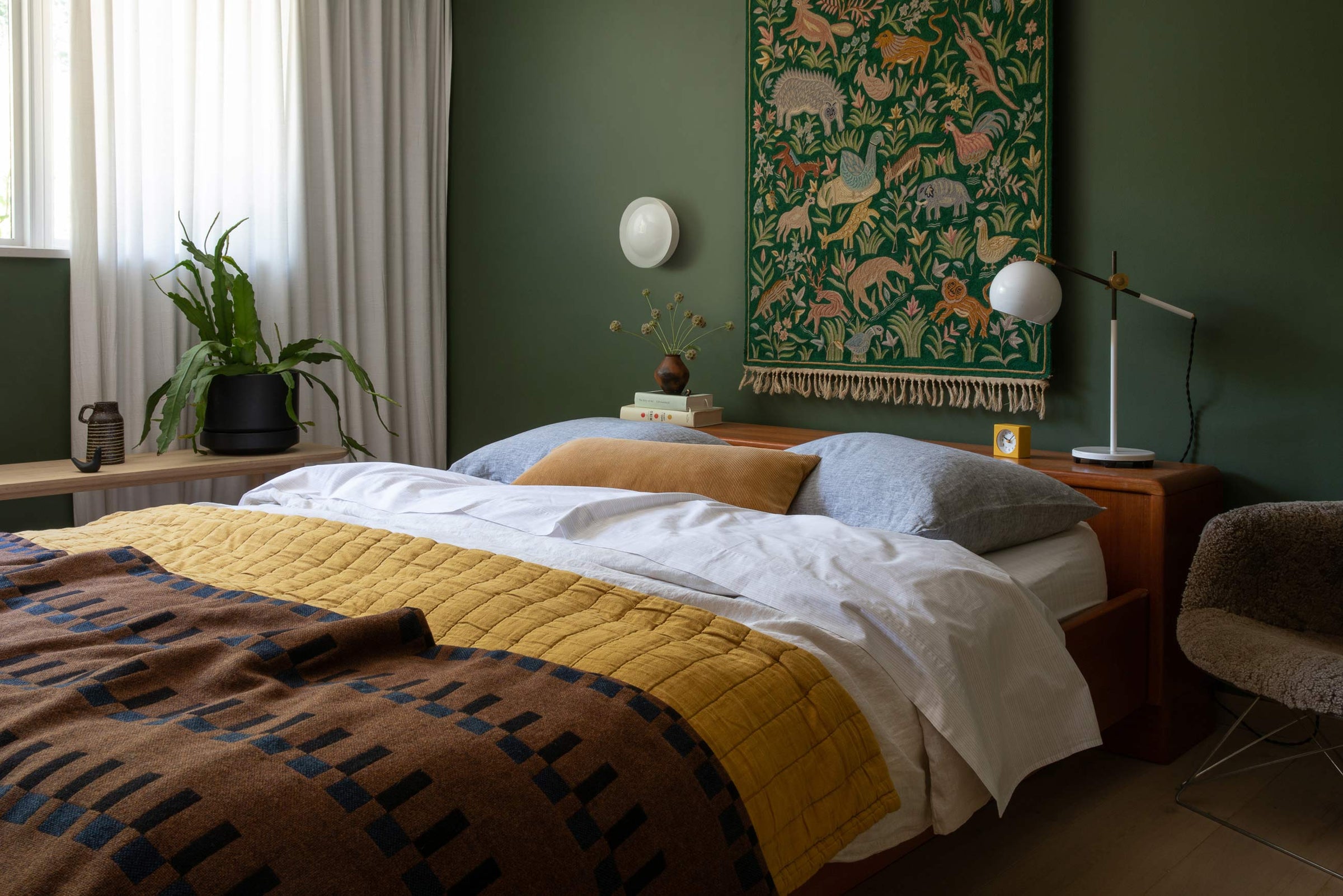 Bed with mustard quilt and patterned throw beneath a white round wall sconce on a deep green wall, wooden nightstand with lamp and tapestry