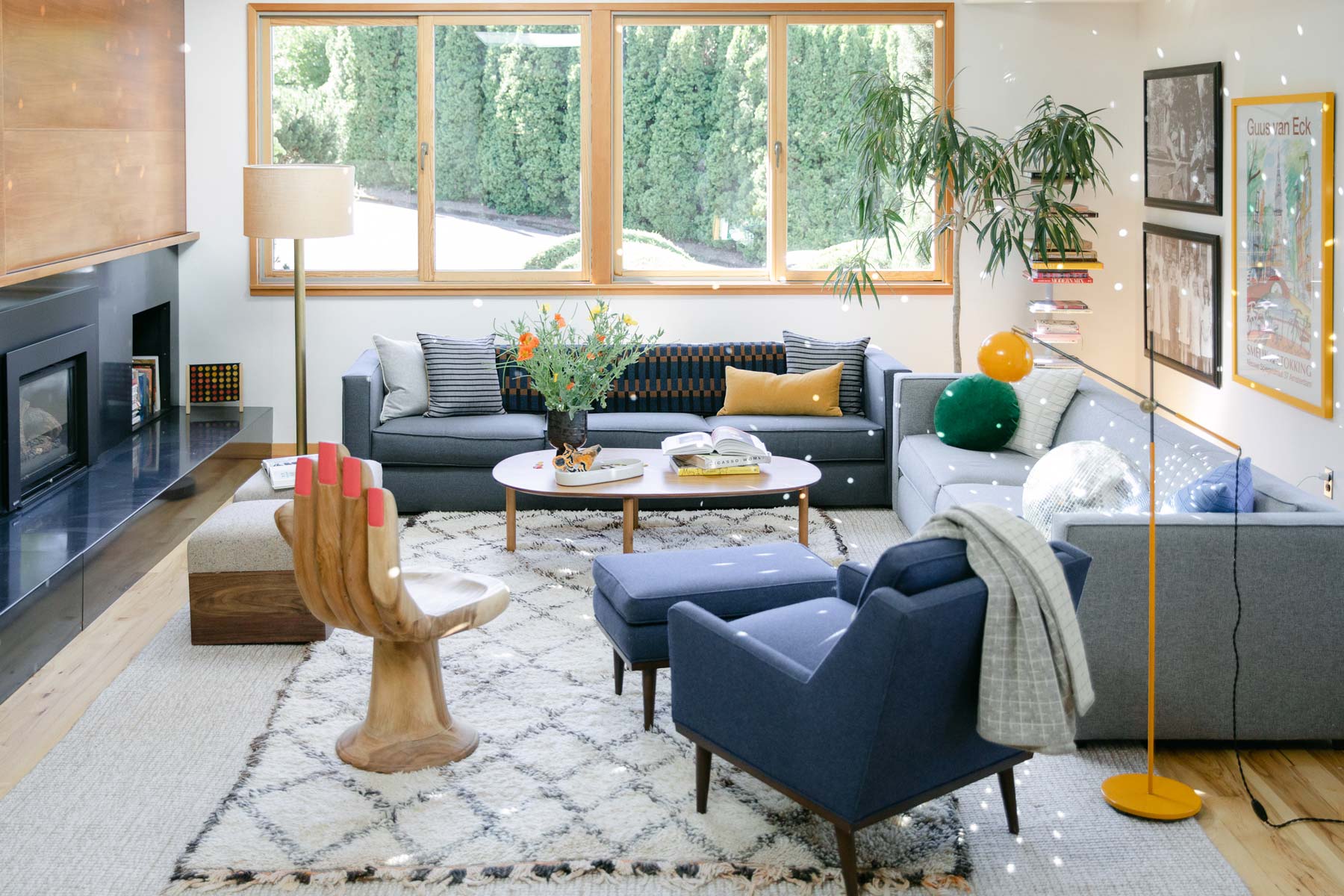Sunlit living room with gray sectional sofa, blue armchair, wooden hand-shaped chair, patterned rug and coffee table with flowers by large window