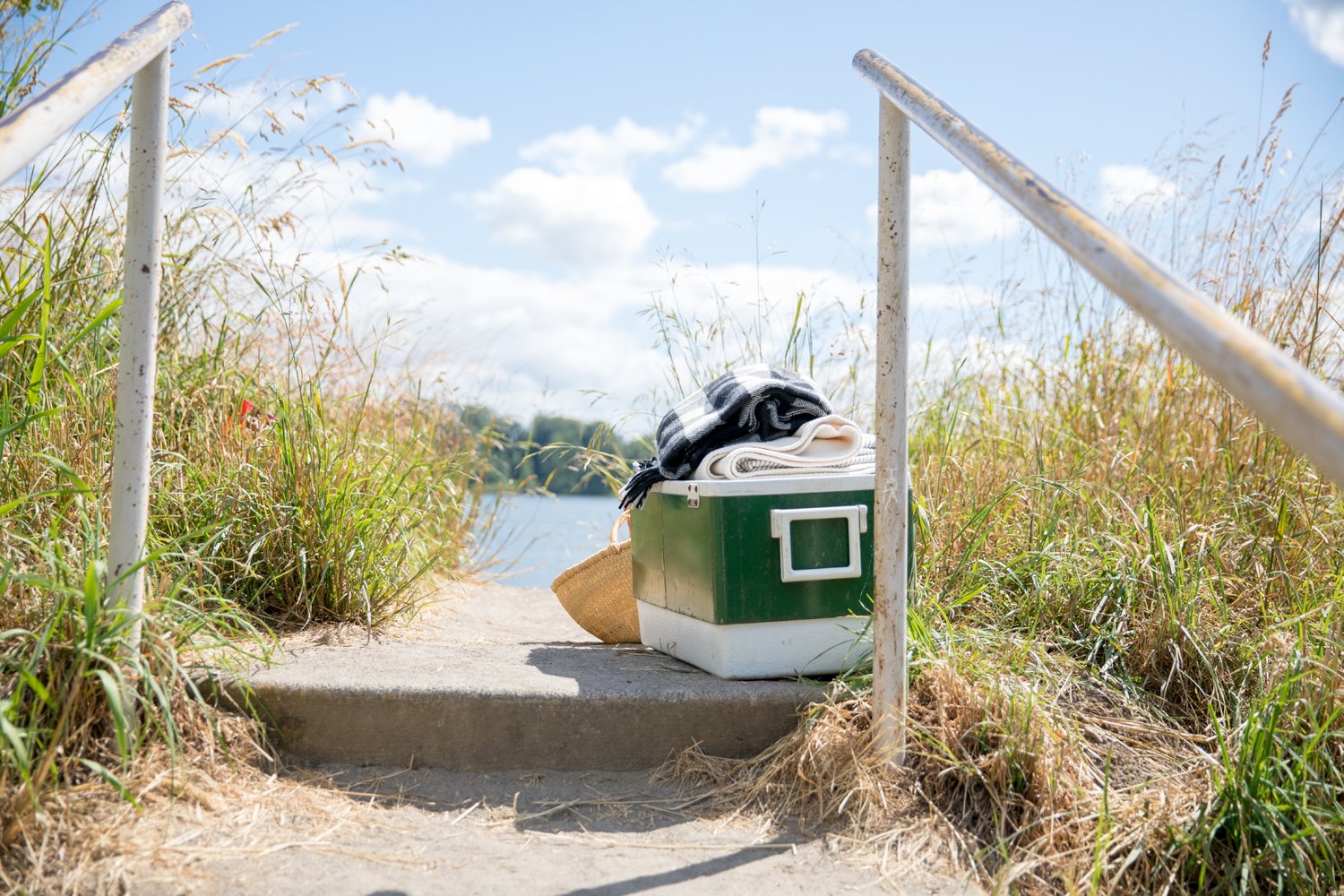 Green cooler and straw hat sitting on a concrete path overlooking a lake, framed by tall grasses and a blue sky
