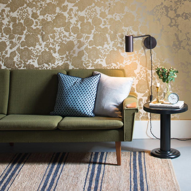 Green mid-century sofa against floral gold wallpaper with wall sconce and black side table holding books a clock and flowers