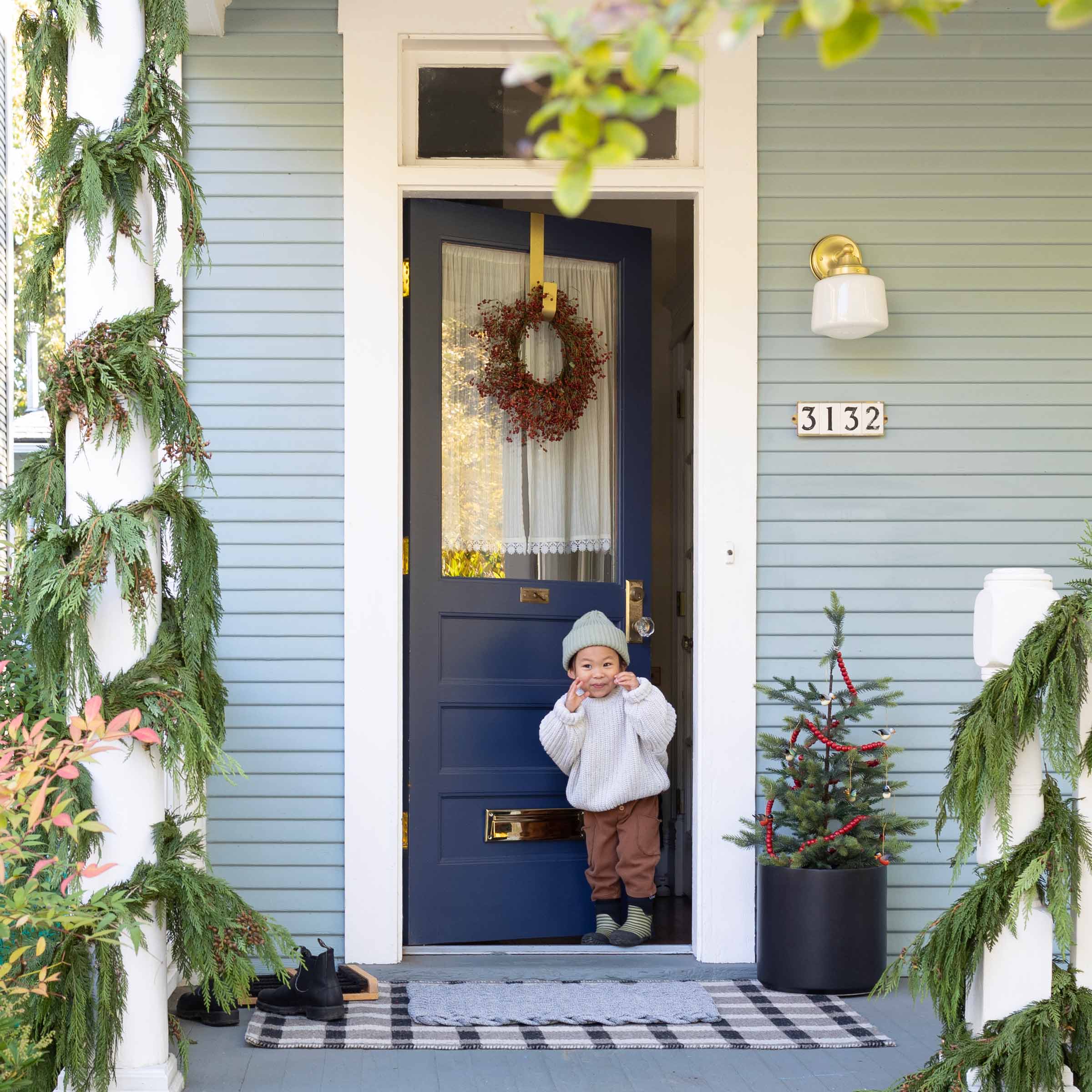 A toddler in front of a home with a red berry wreath and mini tree.