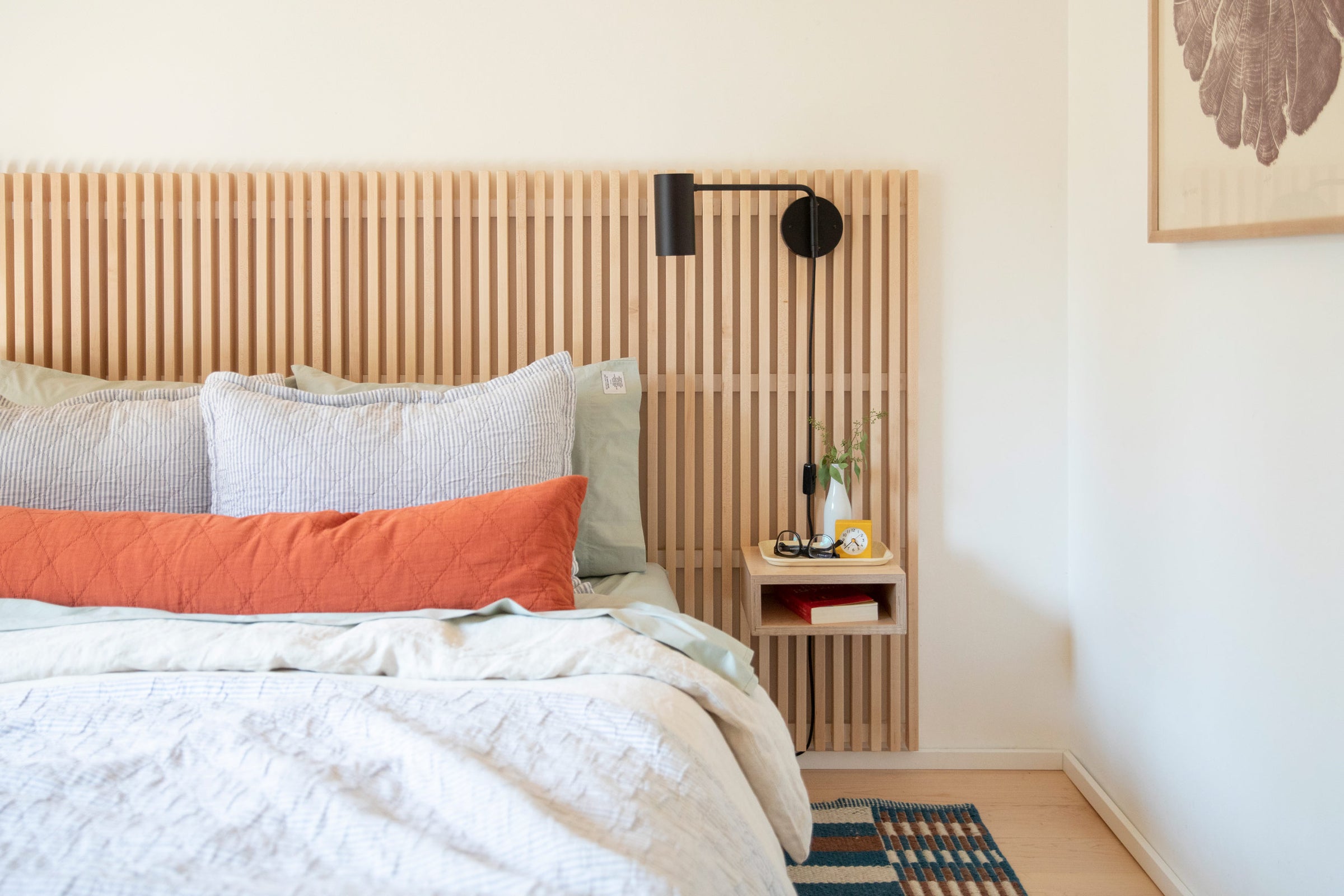 Serene bedroom with light wood slat wall and cozy bed with pinstripe pillows, red bolster pillow, and white duvet next to bedside table with eye glasses, small yellow clock, and bud vase